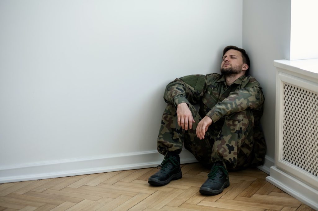 Depressed army man in uniform sitting in a corner of an empty ro